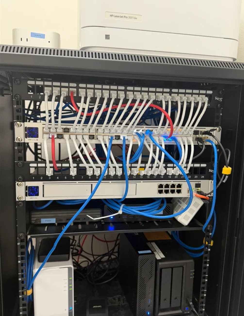 A finished server rack. White network cables are neatly plugged into two rows of patch panels and lead to a switch. Below, a virtualized server and storage sit on the shelves, and a printer and a small computer rest on top.