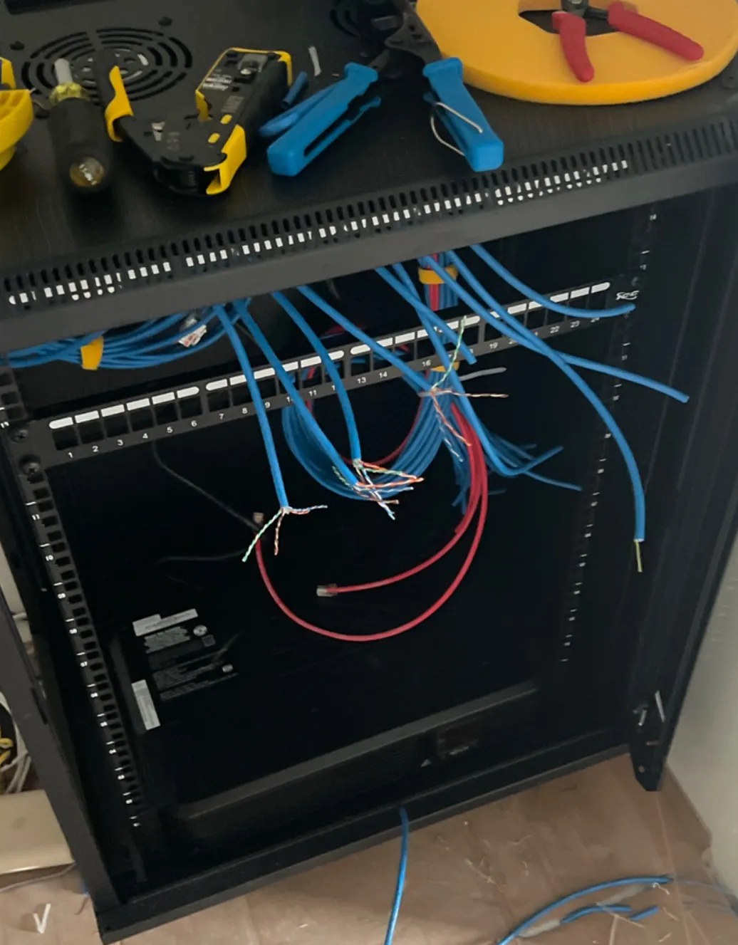 A black server rack with LAN tools, wire cutters, and a cable reel on top. Dozens of blue network cables and a red one hang loose inside the rack, still unsorted during construction.
