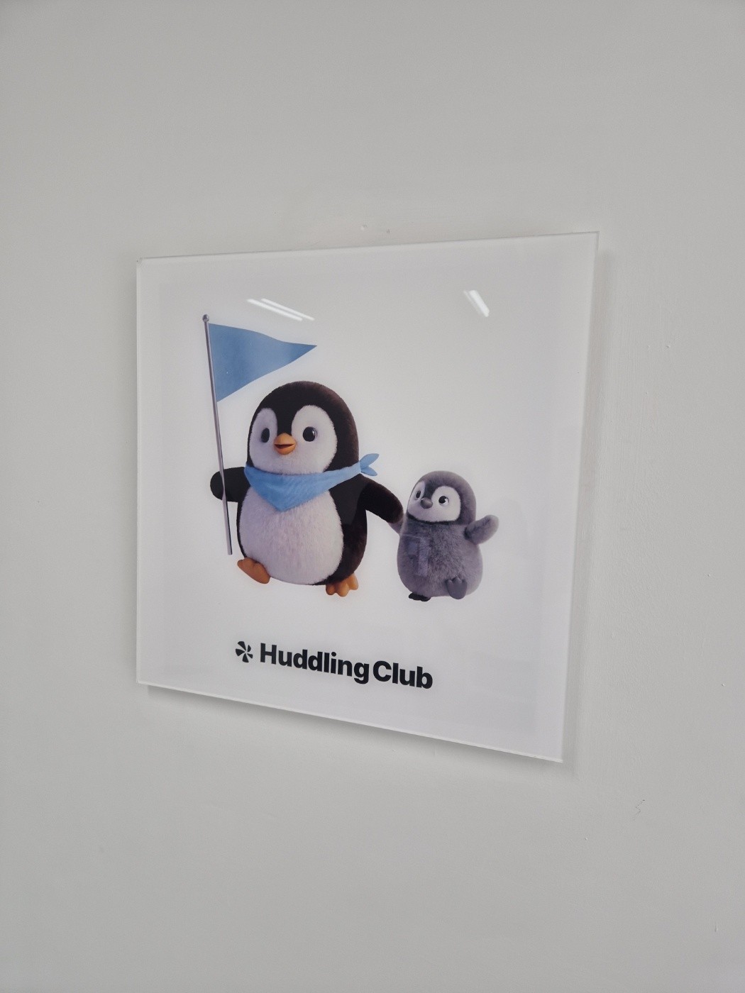 A Huddling Club sign hanging on a wall, showing a penguin holding a flag and a small penguin, with the Huddling Club logo below the drawing