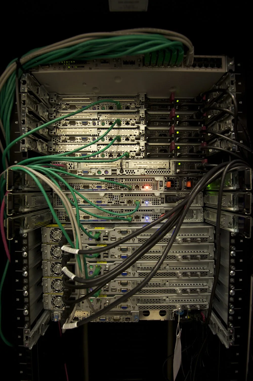 Back of a data center server rack. Each numbered shelf holds several servers side by side, and green and black cable bundles and blinking status lights are tangled together